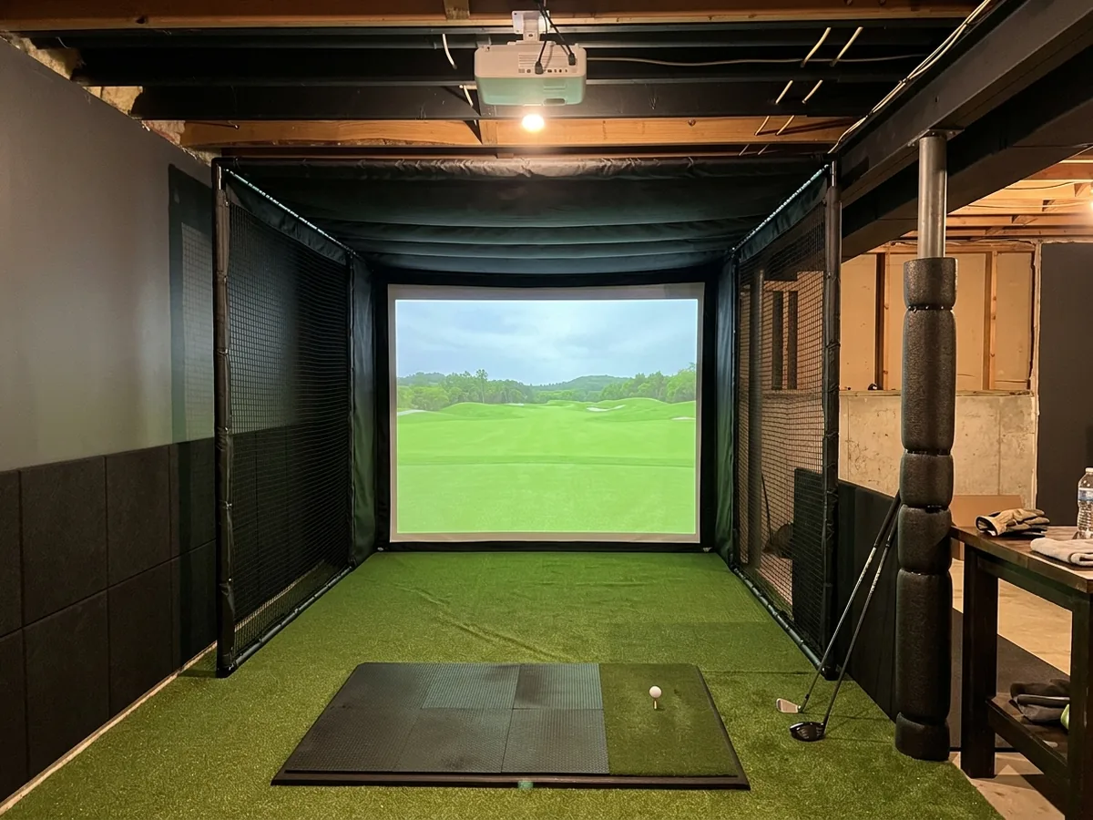 Basement golf simulator bay with a black fabric enclosure, a white impact screen, padded side walls and a hitting mat